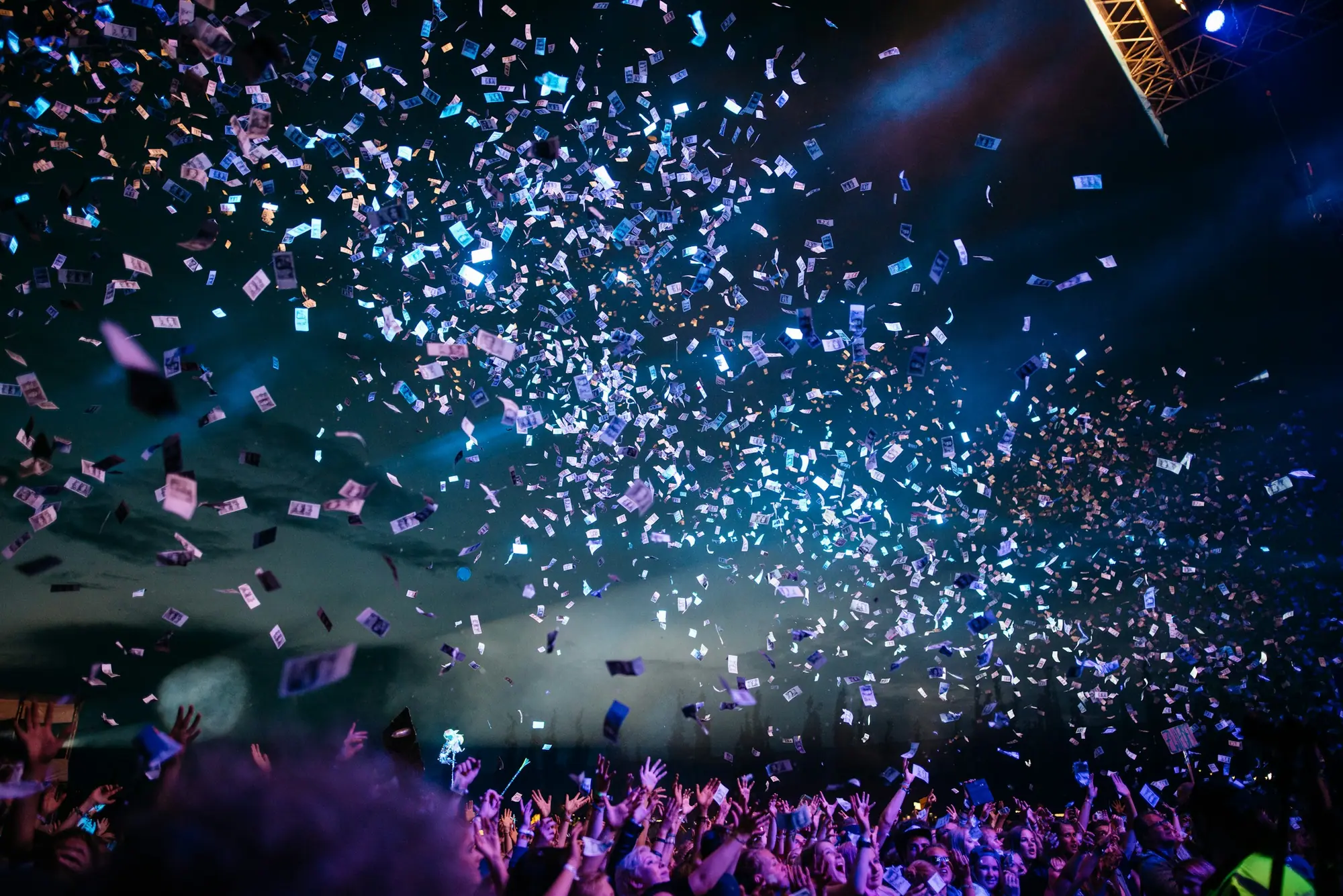 A festival crowd with hands raised as confetti falls under blue stage lights