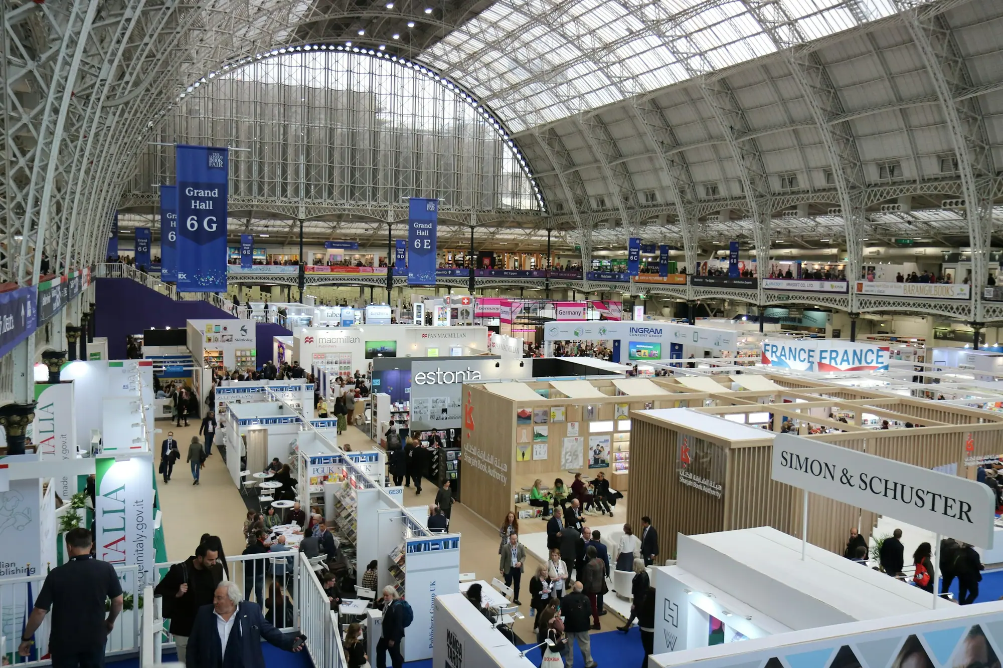 A busy exhibition hall under a glass roof, lined with branded booths